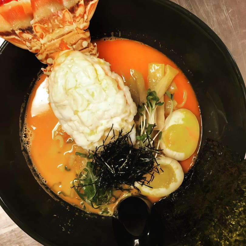Lobster ramen with creamy topping, soft-boiled eggs, seaweed, and sprouts in a black bowl.